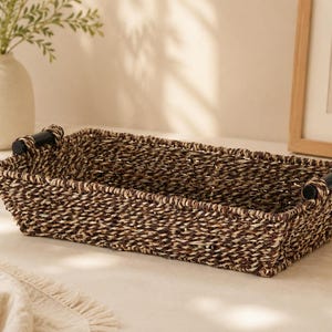 May include: A rectangular woven basket with a dark brown and tan textured finish. The basket features two black cylindrical handles attached to the short sides, providing a sturdy grip for this open-top storage container.