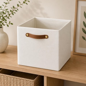 May include: A square, off-white fabric storage bin featuring a single brown faux leather handle attached with two gold-toned metal rivets on the front panel. The container has a structured, open-top design with clean, straight edges and a smooth texture.