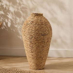 May include: A tall, floor-standing vase crafted from tightly coiled, light brown natural seagrass fibers. The vessel features a tapered, teardrop-like silhouette with a narrow neck and a wide, rounded body, showcasing a textured, woven surface.