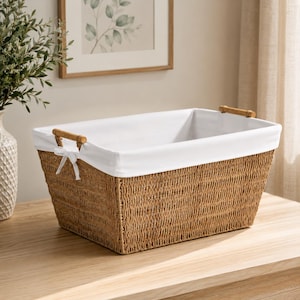 May include: A rectangular woven storage basket made of natural tan fibers. The basket features a removable white fabric liner that folds over the top edges, secured by two wooden handles and a small white ribbon bow on one side of the rim.