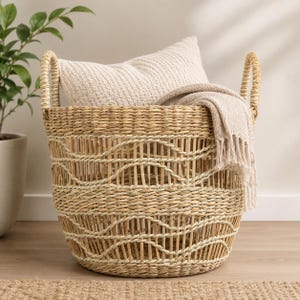 May include: A round woven seagrass basket with two handles, featuring an open-weave pattern with horizontal wavy lines. Inside the basket sits a beige textured throw pillow and a matching beige fringed throw blanket draped over the side.