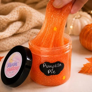 May include: A clear plastic jar filled with orange textured slime, featuring a black label with the white text Pumpkin Pie. The black lid displays a circular sticker with a crescent moon and the text Moon Mellow Slime. Small yellow star-shaped charms are embedded within the orange slime.