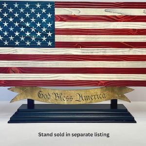 May include: A rectangular wooden American flag with red, white, and blue painted stripes and white stars on a blue field. The flag is mounted on a dark blue rectangular base featuring a gold-colored banner with the text God Bless America.