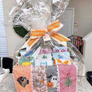 May include: A gift basket wrapped in clear cellophane with a Tennessee ribbon, containing multiple folded fabric towels featuring various seasonal and holiday embroidered designs including a dog, turkey, pumpkin, ghost, and Christmas tree.