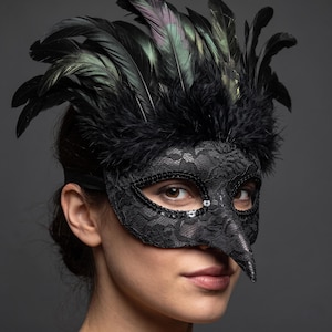 May include: A black masquerade mask featuring a pointed beak-like nose, intricate lace detailing, and a border of black sequins around the eye openings. The top of the mask is adorned with a dense arrangement of iridescent black and green feathers.