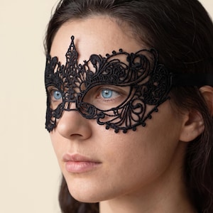 May include: A black lace masquerade mask featuring intricate, swirling floral patterns and a pointed centre detail. The mask covers the eye area and is secured with a thin black ribbon strap that extends around the sides of the face.
