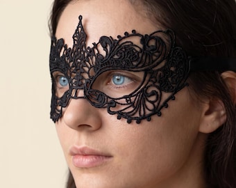 May include: A black lace masquerade mask featuring intricate, swirling floral patterns and a pointed center detail. The mask covers the eye area and is secured with a thin black ribbon strap that extends around the sides of the face.