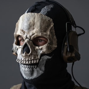 May include: A black balaclava featuring a realistic, textured off-white skull print covering the face area. The mask is worn with tan-colored tactical communication headphones that feature an adjustable microphone boom and a black connecting cable.