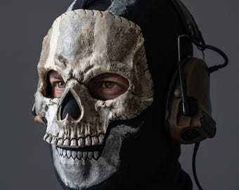 May include: A black balaclava featuring a realistic, textured off-white skull print covering the face area. The mask is worn with tan-colored tactical communication headphones that feature an adjustable microphone boom and a black connecting cable.