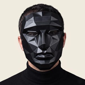 以下が含まれることがあります： 鋭い角張った面を持つ幾何学的なファセットデザインのマットブラックのフルフェイスマスク。マスクには、細かいダークメッシュで覆われた水平な目の開口部と、明確な鼻と唇の構造があり、伸縮性のあるストラップで固定されています。
