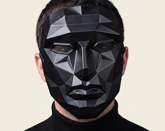 May include: A matte black full-face mask featuring a geometric, faceted design with sharp angular planes. The mask includes horizontal eye openings covered with a fine dark mesh screen and a defined nose and lip structure, secured by an elastic strap.