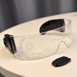 Op de afbeelding: Transparante vizierbril met een zwart montuur en zwarte pootuiteinden. De doorzichtige lens is bedrukt met witte grafische details, waaronder een cirkelvormig doelwit, lineaire circuitpatronen en een liniaalachtige schaalverdeling aan de bovenrand.