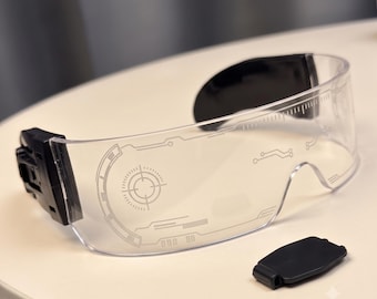 May include: Clear visor glasses featuring a black frame and black temple tips. The transparent lens is printed with white graphic details, including a circular target reticle, linear circuit patterns, and a ruler-like scale along the top edge.
