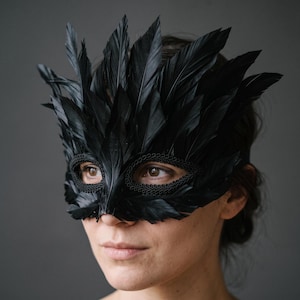May include: A black masquerade mask adorned with layered black feathers extending upwards and outwards. The eye openings are framed with a decorative black braided trim, creating a textured border around the eyes while covering the upper face.