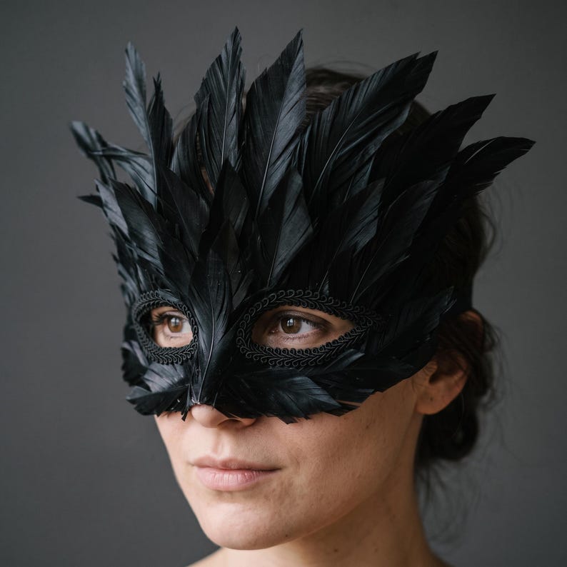 May include: A black masquerade mask adorned with layered black feathers extending upwards and outwards. The eye openings are framed with a decorative black braided trim, creating a textured border around the eyes while covering the upper face.