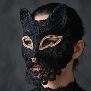 May include: A black lace cat mask featuring pointed ears and intricate floral embroidery patterns. The eye openings and the nose area are embellished with rows of small, sparkling gold-toned rhinestones, creating a defined feline facial structure.