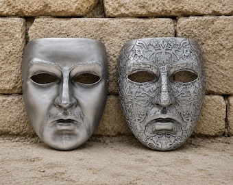 May include: Two silver-colored face masks. The mask on the left features a smooth, realistic human face with subtle sculpted eyebrows and facial hair. The mask on the right displays an intricate, embossed floral and vine pattern across the entire surface.