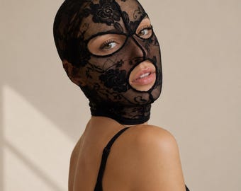May include: A black balaclava crafted from sheer lace fabric featuring an intricate floral pattern. The head covering includes cutouts for the eyes and mouth, fitting snugly over the head and neck, paired with a simple black garment strap.
