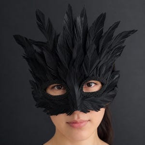 May include: A black mask covered in layered feathers that extend upwards and outwards. The eye openings are framed with a decorative braided trim, creating a textured appearance. The mask covers the upper portion of the face, featuring a pointed design.