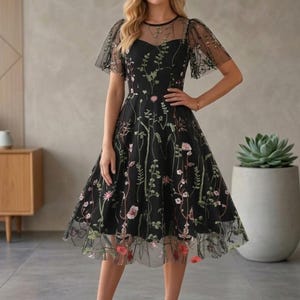 May include: A black knee-length dress featuring a sheer mesh overlay with intricate floral and vine embroidery in shades of green, pink, and white. The garment has short flutter sleeves, a round neckline, and a fitted bodice with a flared skirt.