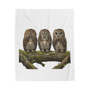 May include: A white fleece blanket featuring a graphic illustration of three brown owls perched on a mossy tree branch. The first owl covers its eyes with its wings, the second owl has its eyes closed, and the third owl covers its beak.