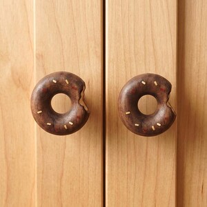 May include: Two dark brown wooden cabinet knobs shaped like donuts with a bite taken out of the side. The knobs feature small, inlaid light-colored and red rectangular accents that resemble sprinkles on the surface of the dark wood.
