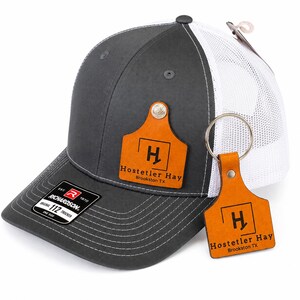 以下が含まれることがあります： チャコールグレーと白のメッシュトラッカーキャップ。家畜用耳標の形をしたタンカラーのレザーパッチには、黒い文字で「Hostetler Hay Brookston TX」と記されています。同じロゴが入ったレザー製のキーホルダーがキャップの側面に付いています。