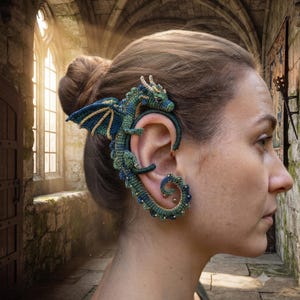 May include: A detailed crochet ear cuff shaped like a dragon, featuring shades of teal, blue, and gold thread. The dragon's body wraps around the outer edge of the ear, with its head resting at the top and a curled tail extending toward the earlobe.