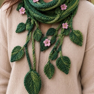 May include: A knitted scarf in various shades of green featuring dangling vine-like strands with textured green leaves and small, five-petaled light pink flowers attached throughout the length of the accessory, worn over a beige knit sweater.