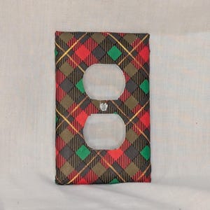 May include: A rectangular electrical outlet wall plate cover featuring a diagonal plaid pattern in shades of red, dark green, black, and thin gold lines. The cover has two circular openings for a duplex receptacle and a center screw hole.