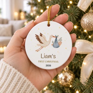 Könnte beinhalten: Ein rundes, weißes Keramikornament mit einer Aquarellillustration eines Storchs, der ein blaues Bündel mit einem Baby trägt. Der Text lautet Liam's FIRST CHRISTMAS 2026 in brauner Schrift. Das Ornament hängt an einer goldenen Schnur.