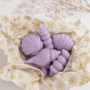 Puede incluir: Una colección de figuras de cera en forma de conchas marinas de color violeta claro, dispuestas sobre papel de seda blanco con un patrón de copos de nieve dorados. El conjunto incluye varias conchas en espiral y en forma de abanico.