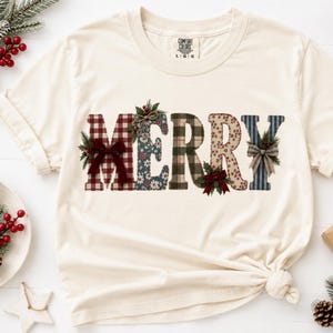 Könnte beinhalten: Ein cremefarbenes Kurzarm-T-Shirt mit dem Wort MERRY in großen, gemusterten Buchstaben. Jeder Buchstabe hat ein einzigartiges Design, darunter rotes Karomuster, Blumenmuster, grünes Karomuster und blaue Streifen, akzentuiert mit kleinen Schleifen.