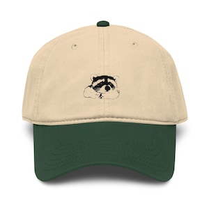 Può includere: Un cappellino da baseball con corona beige e visiera verde scuro. Il pannello frontale presenta una piccola illustrazione grafica in bianco e nero centrata di un procione a riposo con la testa china, resa in uno stile dettagliato a schizzo.