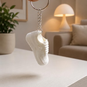 Könnte beinhalten: Ein weißer Schlüsselanhänger in Form eines Sneakers mit detailliertem Miniatur-Schuhdesign, Schnürsenkeln, perforierter Zehenkappe und seitlichem Swoosh, befestigt an einem silberfarbenen Metallring durch eine kurze Gliederkette.
