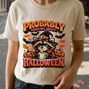 Könnte beinhalten: Ein beiges T-Shirt mit einer Grafik eines Waschbären, der einen Hexenhut trägt und eine Tasse hält. Der Text "PROBABLY THINKING ABOUT HALLOWEEN" steht in orangefarbenen und rosa Buchstaben, umgeben von herbstlichen Blättern, schwarzen Fledermäusen und Kürbissen.
