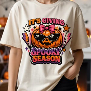 May include: A cream-colored t-shirt featuring a graphic of an orange jack-o'-lantern wearing black sunglasses and a pink bow. The text above and below the pumpkin reads IT'S GIVING SPOOKY SEASON in colorful, bold, retro-style lettering with stars and lightning bolts.