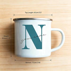 May include: A white enamel mug with a silver rim and a teal letter N printed on the front, accompanied by the text your name. Dimensions are 3.5 inches top length, 3.15 inches height, and 2.95 inches bottom diameter as indicated by labels.