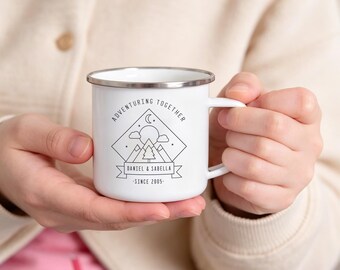 May include: A white enamel mug with a silver rim featuring a black line art graphic of mountains, a sun, clouds, and a crescent moon. The text reads ADVENTURING TOGETHER, DANIEL & SABELLA, and SINCE 2005. The mug has a side handle and a smooth finish.