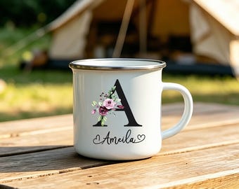 May include: A white enamel mug with a silver rim featuring a large black letter A decorated with a cluster of pink and dark purple flowers. Below the letter, the name Ameila is printed in black script, flanked by two small black heart icons.