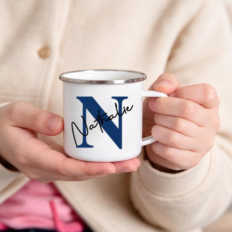 May include: A white enamel mug with a silver rim and a handle. The mug features a large navy blue letter N printed on the front, with the name Nathalie written in black cursive script layered over the letter. The mug has a smooth, glossy finish.