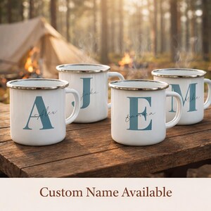 May include: Four white enamel mugs with silver rims, each featuring a large blue letter and a custom name in script font. The mugs are arranged on a wooden surface with steam rising from them. The text Custom Name Available is at the bottom.