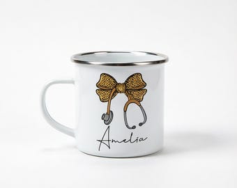 May include: A white enamel mug with a silver rim featuring a graphic of a leopard print bow attached to a stethoscope. The name Amelia is printed in black cursive script below the stethoscope graphic on the front of the mug.