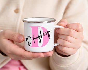 May include: A white enamel mug with a silver rim and a handle, featuring a large pink letter D printed on the front. Overlaid on the pink letter D is the name Deloughery written in a black cursive script font across the center of the mug.