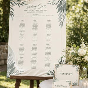 Könnte beinhalten: Ein großer Sitzplan für eine Hochzeit auf einer Holzstaffelei mit dem Text Seating Chart, Emma & James, 14th June 2025 sowie einer Liste von Tischen und Gästen. Das Schild hat grüne Blattmotive in den Ecken und einen cremefarbenen Hintergrund.