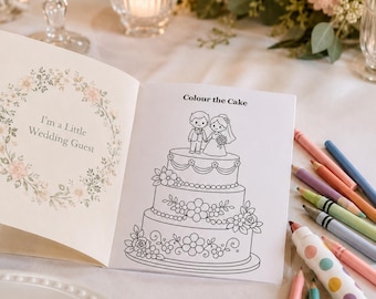 Puede incluir: Un libro para colorear abierto titulado I'm a Little Wedding Guest con una página que presenta una ilustración de un pastel de bodas de tres niveles y el texto Colour the Cake, acompañado de varios lápices de colores en tonos rosa, azul y verde.