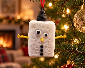 Puede incluir: Un adorno navideño blanco con forma de malvavisco rectangular, con sombrero negro, brazos amarillos, ojos móviles, nariz amarilla y tres botones negros, colgado de una cinta roja en un árbol de pino verde con luces.
