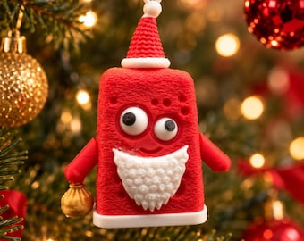 Puede incluir: Un adorno navideño rectangular rojo con forma de personaje que lleva un gorro de Papá Noel y una barba blanca. La figura tiene dos ojos grandes móviles, brazos rojos pequeños y sostiene un pequeño saco dorado en una mano.