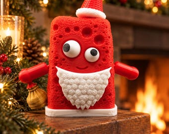 Puede incluir: Un personaje de esponja rectangular rojo con textura porosa, que presenta grandes ojos saltones, una barba blanca rizada y un gorro de Papá Noel rojo. La figura tiene brazos rojos, sostiene un pequeño saco dorado y tiene un borde blanco en la base.