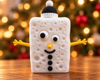 Puede incluir: Una figura rectangular de esponja blanca con textura porosa, que presenta dos ojos móviles, una pequeña nariz amarilla, una boca negra curvada y tres botones negros. Tiene brazos de plástico amarillo y un gorro negro tejido.